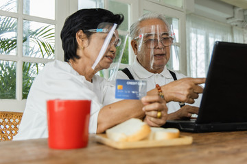Portrait of an elderly person in Asia shopping online via a laptop. They pay via credit card. Retirees wear face shields for daily activities. new normal concept