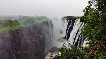 The unique natural wonder of Victoria Falls. In a gorge with steep rocky slopes, water flows from the Zambezi River. Over the waterfall there is a thick fog of water spray. Green vegetation around. 
