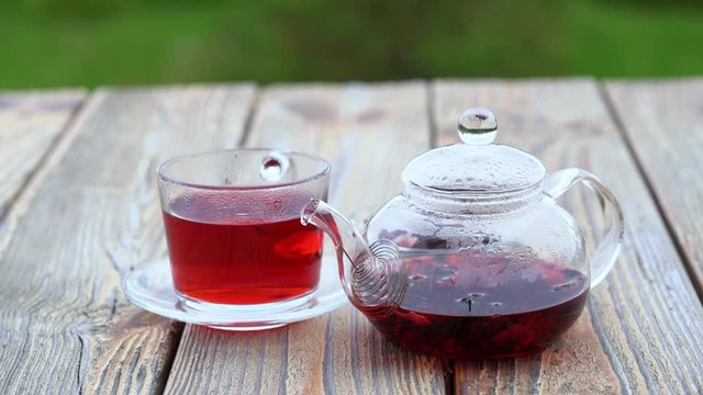 A transparent teapot and a glass cup of hibiscus karkade herbal tea on a wooden table, outdoors on green blurred background.