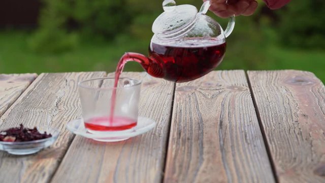 The process of pouring hibiscus karkade herbal tea from glass teapot into a Cup, outdoors on the terrace on green blurred background.