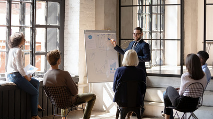 Smiling businessman coach giving flip chart presentation at meeting, pointing at diagrams, mentor...