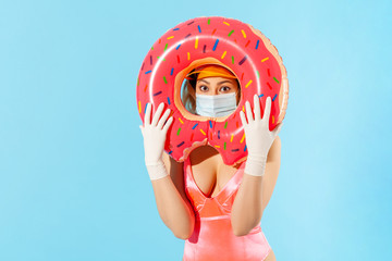 Woman in swimsuit looking through rubber ring, wearing hygienic face mask and surgical gloves to prevent contagious coronavirus on resort beach, rest and summer vacation with covid-19. studio shot