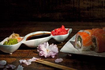 Sushi on a white plate. Sushi roll with sauce and spices on a black background. Food on a wooden table with dark boards. Sakura color on a plate. Flower with rose petals.