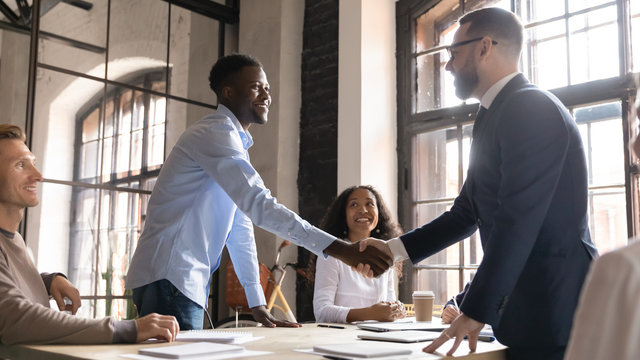 Happy diverse business partners handshaking at successful group negotiation, making agreement, good legal deal, signing contract, African American and Caucasian businessmen shaking hands at meeting