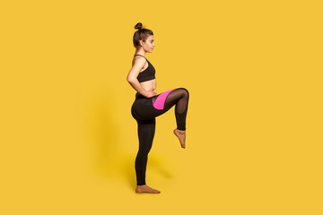 Fototapeta na wymiar Side view, athletic slim woman with hair bun in tight sportswear doing sport standing with one leg lift, thighs workout, warming up training muscles. full length studio shot, isolated on yellow