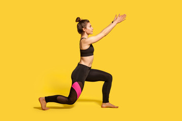 Fototapeta na wymiar Side view, slim young woman with hair bun in tight sportswear doing sport lunge exercise, standing one knee and raising hands, warming up training muscles. full length studio shot, isolated on yellow