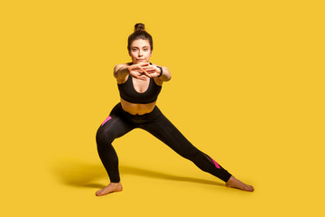 Fototapeta na wymiar Athletic pretty girl with hair bun in tight sportswear doing sport, lower-body exercise, stretching hands out, warming up training muscles for flexibility. full length studio shot, isolated on yellow
