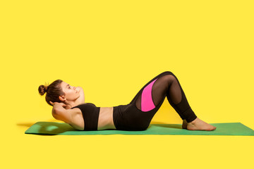 Fitness young woman with hair bun in tight sportswear training on gym mat, doing sit ups, exercising abdominal muscles, lower-body workouts, warming up. studio shot, isolated on yellow background - obrazy, fototapety, plakaty