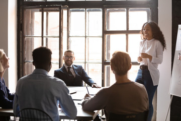 Smiling African American business coach laughing at joke at briefing, giving flip chart presentation, diverse colleagues having fun at corporate meeting in boardroom, good team relationship