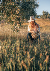 Obraz premium Happy young girl with a basket in a field of wheat and olive trees with a hat on a sunset. Summer, rural vacation and nature concept.