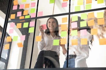 Two smiling diverse businesswomen planning project on sticky papers, adding colorful post it notes with tasks or startup ideas on glass wall in office, coaches working with scrum board together