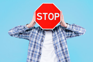 Man is holding red prohibition road sign stop in front of face on blue background. Guy is warning about danger, risk. Prevention symbol, attention icon that shows traffic ban. Caution, alert concept. - obrazy, fototapety, plakaty
