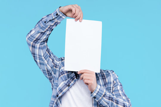 Man In White T-shirt, Checkered Shirt Is Holding Blank Empty Paper Canvas In Front Of Face. Guy Student Is Standing On Blue Background. Template For Advertising, Greeting, Invitation Card Concept.