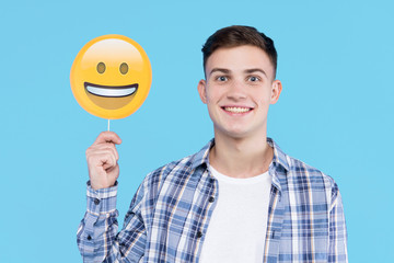 Young man in white t-shirt, checkered shirt is holding in hand paper decorative emoticon, emoji...