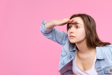 Pretty pensive girl student dressed in white t-shirt, denim jacket is standing on pink background. Young woman is looking, peering to left side. Female gesturing. Emotional portrait concept.