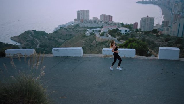 Determined to get fit and healthy, young woman in workout clothes runs up mountain, looks over city skyline at sunrise or sunset. Female athlete during hard rewarding training on road to success