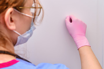 A female doctor in a medical mask knocks on the door of a patient with a coronavirus. Close-up of a nurse in protective gloves near the door of a house