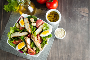 Fresh Caesar salad made of tomato, ruccola, chicken breast, eggs, arugula, crackers and spices. Organic ingredients in a white, transparent bowl on wooden background