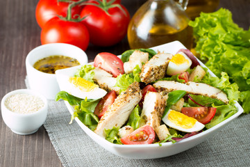 Fresh Caesar salad made of tomato, ruccola, chicken breast, eggs, arugula, crackers and spices. Organic ingredients in a white, transparent bowl on wooden background