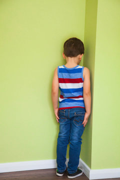 a young boy in a striped t-shirt stands in the corner with his face turned away because he has been punished