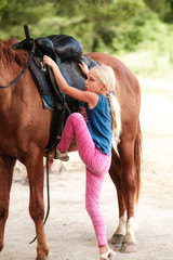 Cute young girl riding a brown horse on a country road at sunset. The child quietly travels on a stallion. The girl confidently controls the horse in a blooming field. Children's summer sports camp.