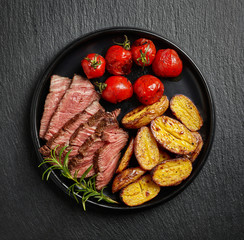 Grilled beef fillet steak with potatoes and tomatoes on black plate on black background, top view