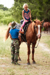 The instructor helps the girl to saddle a brown horse in the forest. Children's equestrian camp....