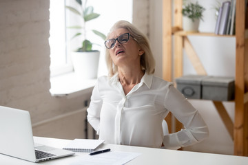 Unhealthy middle-aged female employee sit at desk in office suffer from lower back strain or spasm...