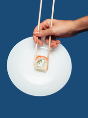 Female hand takes chopsticks sushi rolls with a white round plate on a blue background.