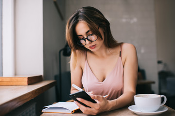 Focused young woman taking notes in daily planner while using smartphone in cafe