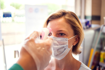 Measuring temperature with digital thermometer in the entrance to market. Woman in medical face mask. Coronavirus and healthcare concept