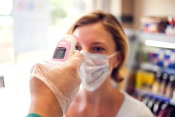 Measuring temperature with digital thermometer in the entrance to market. Woman in medical face...