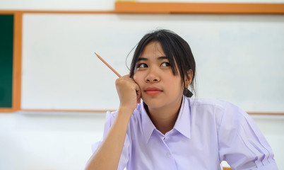 Obraz premium asian student girl thinking for taking homework or reading book alone in classroom with smiling at front green whiteboard, Education learning by myself in white uniform on desk stack of books
