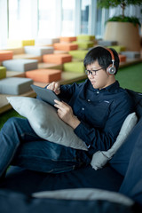 Obraz premium Asian businessman with headphones listening to music working with digital tablet on comfortable sofa at home office. Man with glasses doing online conference on mobile app. Work from home concept