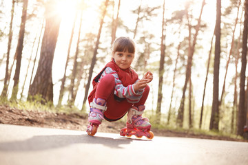 Picture of funny pleasant pretty kid sitting on road in inline skates, looking directly at camera,...