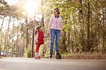 Outside picture of pleasant tender sweet young female having rest with her little daughter, holding her hand, teaching to rollerskate, enjoying time together, being in good mood. Family concept.