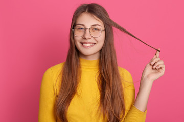 Indoor portrait of adorable girl looking directly at camera, touching her hair, wearing eyeglasses...