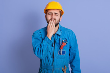 Image of sleepy exhausted builder yawning, covering his mouth with hand, closing eyes, having beard, wearing uniform and helmet, being tired of work, having desire to sleep. People and work concept.