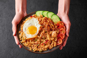 Nasi Goreng - Indonesian Chicken Fried Rice on black plate in female hands. Nasi Goreng is an Indonesian cuisine dish with jasmine rice, chicken meat, fried onion, egg and vegetables. Indoneisan Food