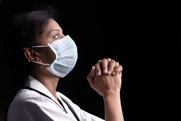Profile view of young woman doctor in medical mask praying to god on black background looking up -...