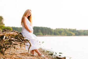 Pregnant blonde woman in long white dress on the beach. Future mother with belly