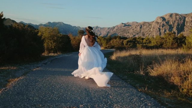 Slow motion woman running at sunset in a white wedding dress on a background of mountains, her hair develops in the wind, freedom and happiness concept
