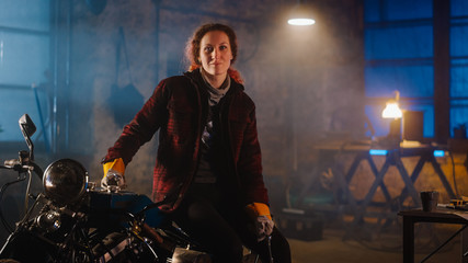 Fototapeta na wymiar Young Authentic Female Mechanic is Sitting on a Custom Bobber Motorbike and Posing in Workwear with Ratchet and Spanner. Talented Girl Wearing a Checkered Shirt. Creative Workshop Garage.