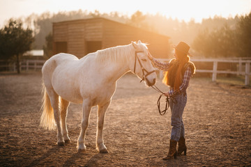 A young cowgirl woman stroking a horse in the sunset at her ranch. Flare effect.