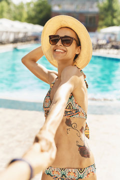 POV and Follow me. Beautiful suntanned smiling woman in hat and sunglasses holding someone hand against swimming pool. Summer vacation, pool lesure