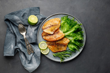 Grilled fish sea bass fillet with green lettuce in a plate on a black stone background. Above view.
