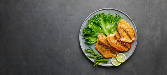 Grilled fish sea bass fillet with green lettuce in a plate on a black stone background. Top view with copy space.