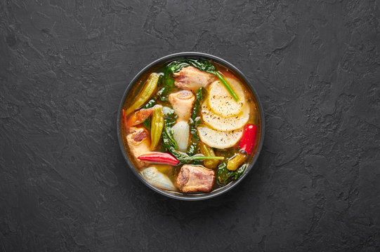 Sinigang na Baboy or Filipino Pork Meat Soup in black bowl on dark slate backdrop. Sinigang is Filipino cuisine dish with meat, bamia, daikon, spinach, fish sauce. Filipino Food. Asian Meal. Top view