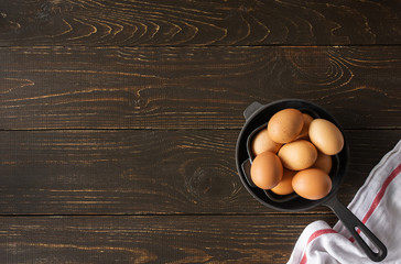 Black pan with fresh raw eggs on brown wooden background, breakfast ingredients, top view, copy...