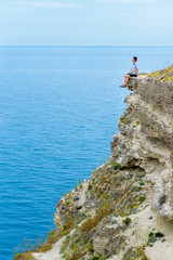 a man sits on the edge of a high cliff and looks out to sea